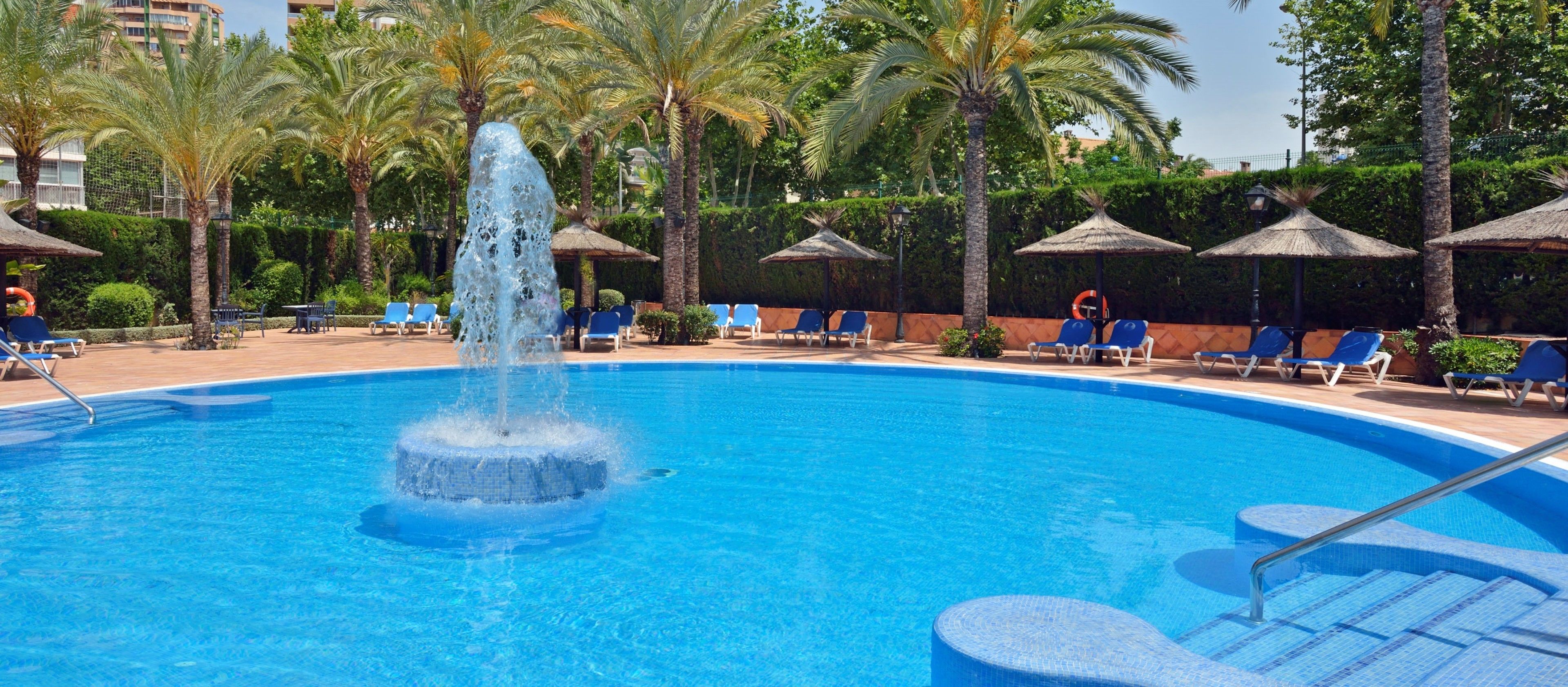 A swimming pool with a central fountain, surrounded by palm trees and lounge chairs.