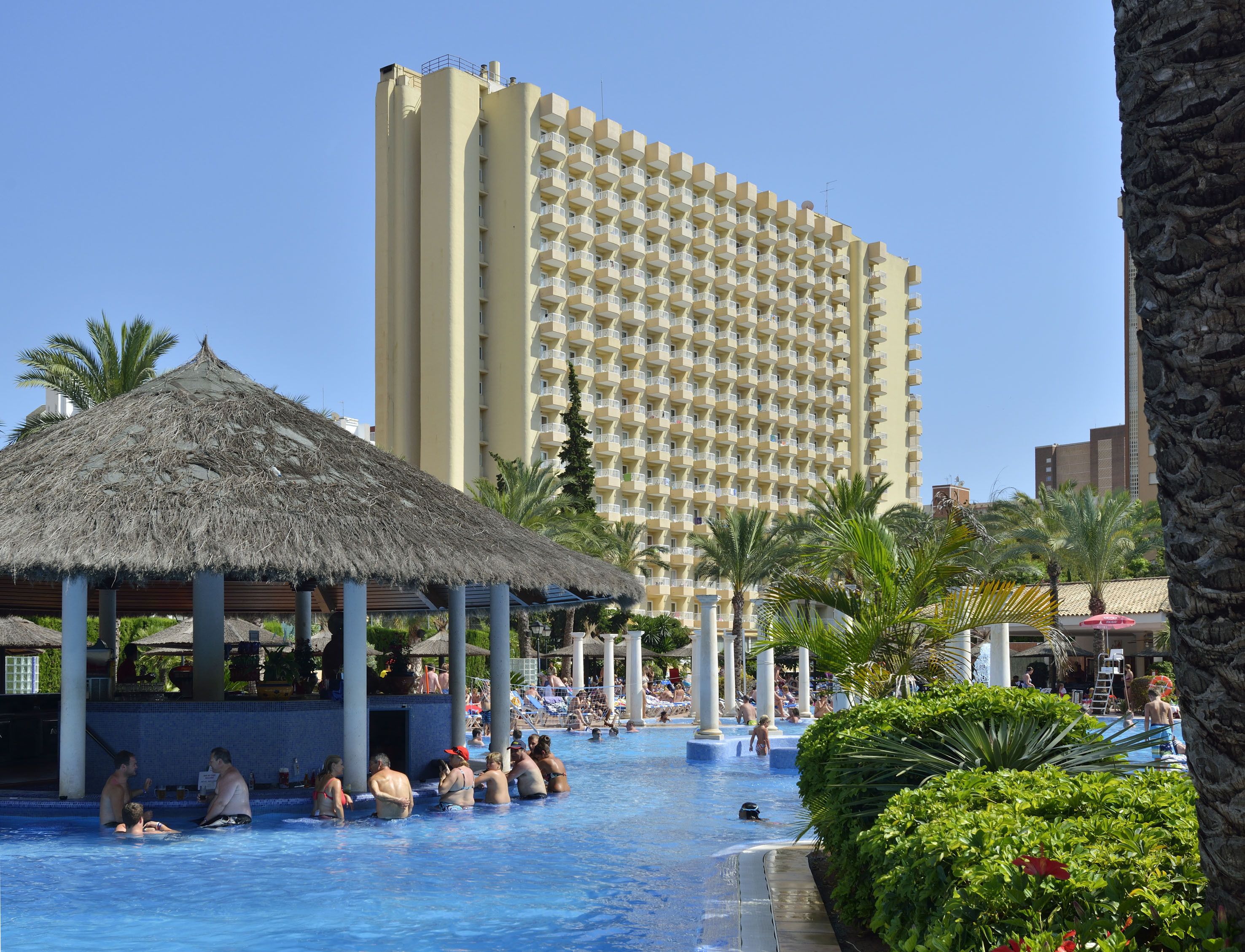A swimming pool with lounge chairs and a thatched-roof structure, surrounded by palm trees and a tall hotel building in the background.