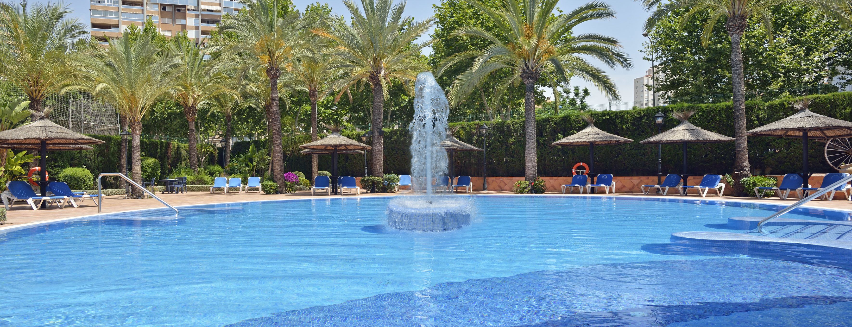 Una piscina con una fuente central, rodeada de sillas de descanso y palmeras.