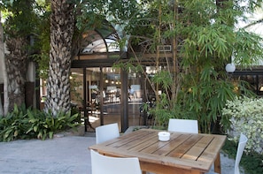 Mesa de madera al aire libre con sillas blancas, rodeada de abundante vegetación y la entrada de un edificio.