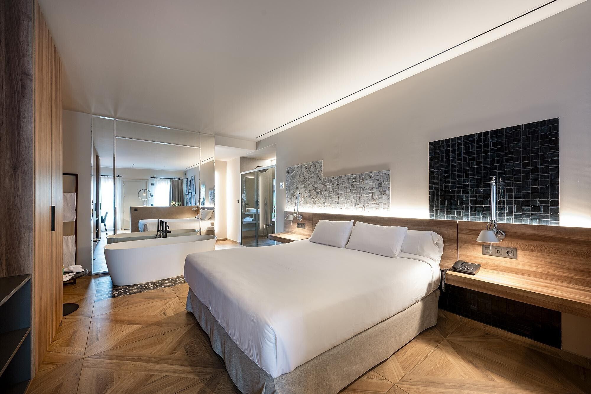 Ein modernes Hotelzimmer mit einem großen Bett, einem Nachttisch mit Lampe, einem Wandbild und einem durch eine Glastrennwand einsehbaren Badezimmer.