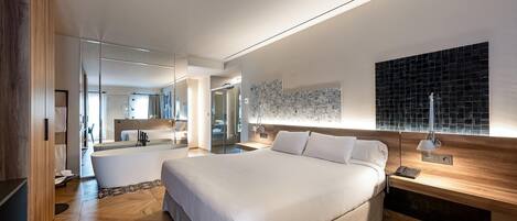 A modern hotel room with a large bed, a bedside table with a lamp, a wall-mounted artwork, and a bathroom visible through a glass partition.