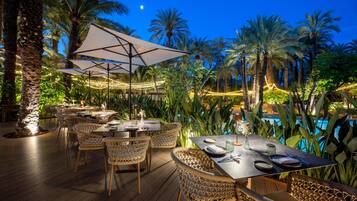 An outdoor dining area with tables and chairs, surrounded by palm trees and a pool.