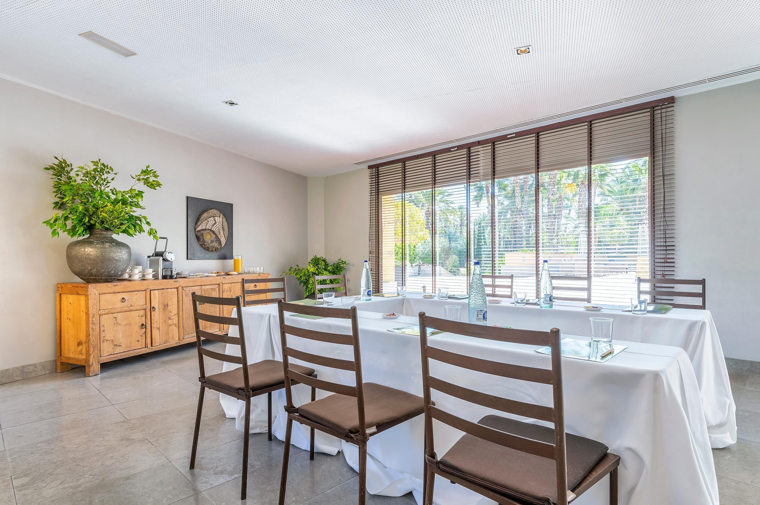 Un comedor con una mesa larga, sillas, un aparador de madera y un jarrón grande con plantas.