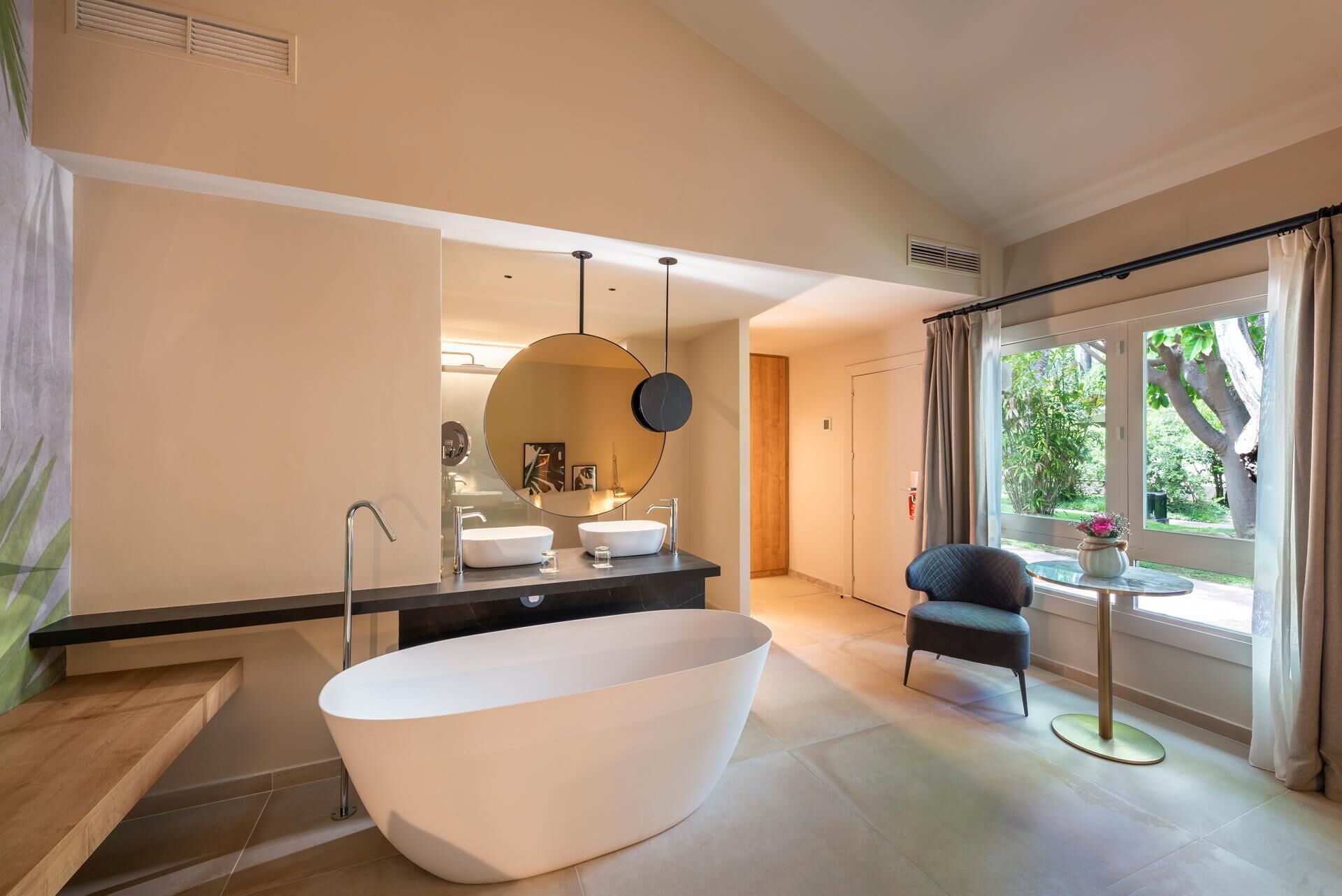 A modern bathroom with a freestanding bathtub, a large mirror, and a vanity with two sinks. There is a chair and a small table by the window, which offers a view of greenery outside.