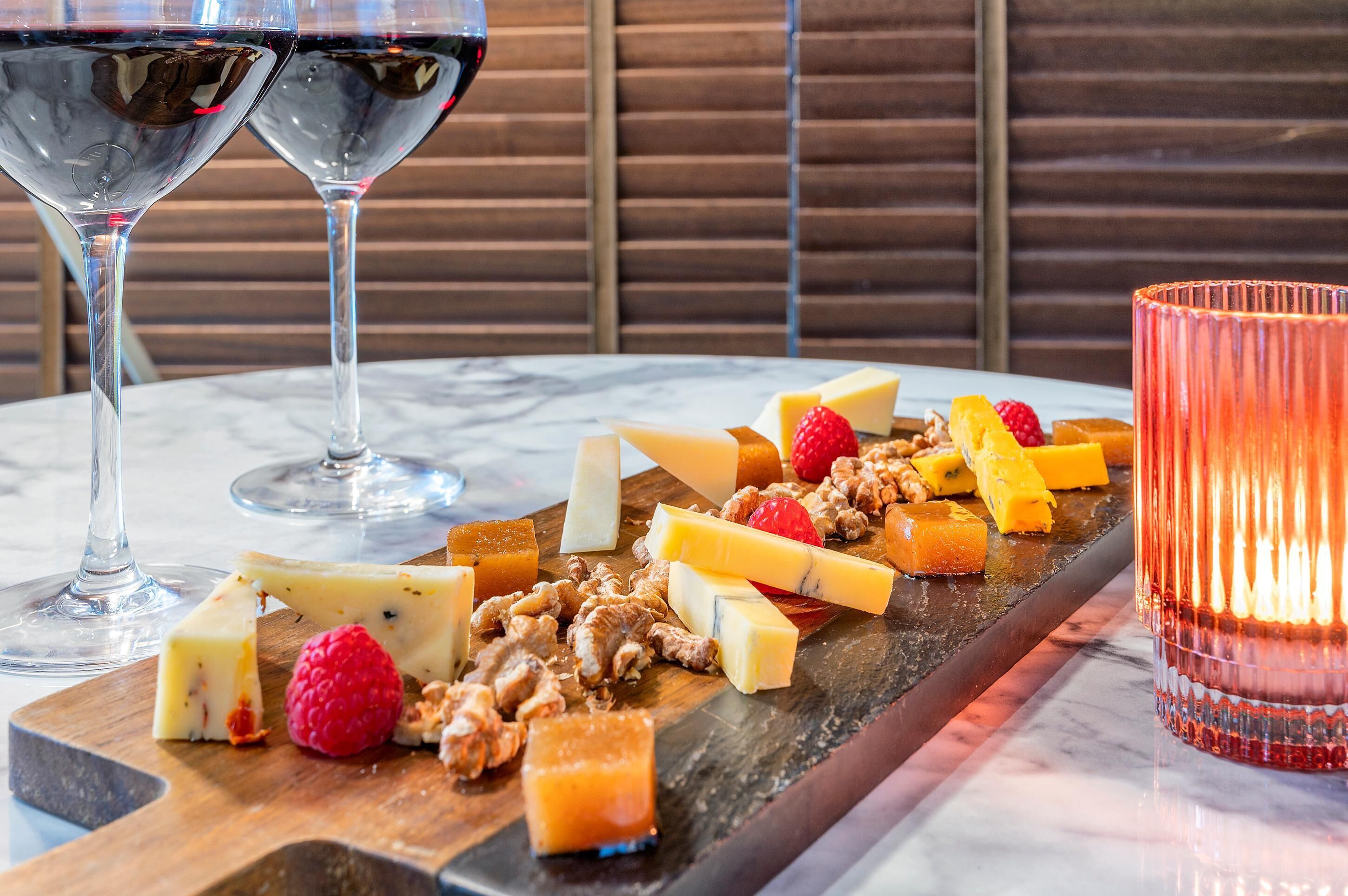 Una mesa de mármol con una tabla de madera que muestra diferentes quesos, frutas y frutos secos. Dos copas de vino tinto están colocadas a cada lado de la tabla.
