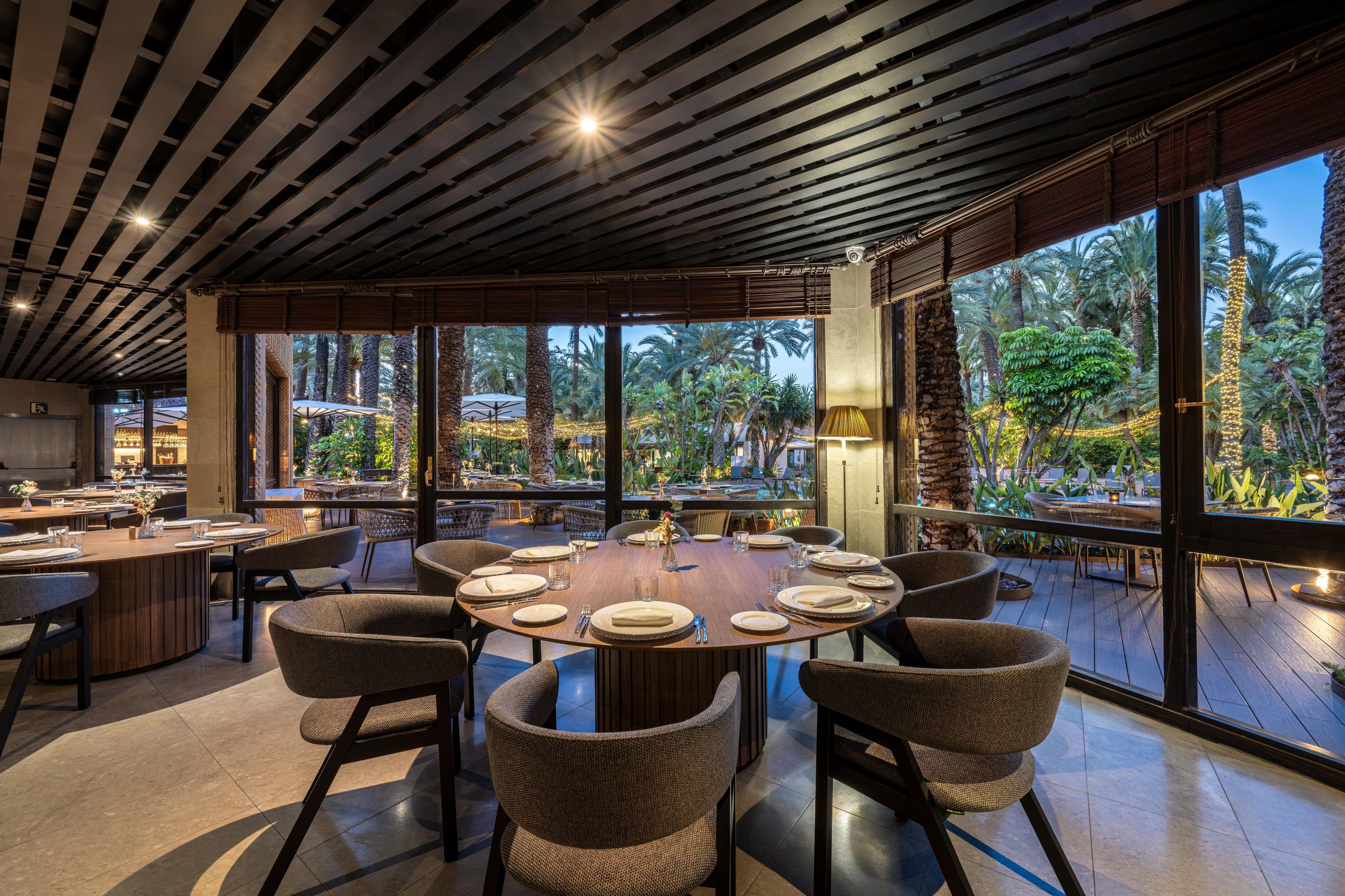 A spacious dining area with a round table set for six, surrounded by wicker chairs, large windows offering a view of palm trees, and a ceiling with wooden slats.