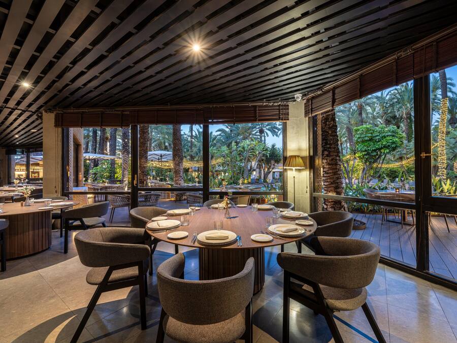 Un comedor amplio con una mesa redonda preparada para seis personas, rodeada de sillas de mimbre, ventanas grandes que ofrecen vista a palmeras y un techo con listones de madera.
