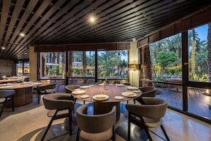 Un comedor amplio con una mesa redonda preparada para seis personas, rodeada de sillas de mimbre, ventanas grandes que ofrecen vista a palmeras y un techo con listones de madera.
