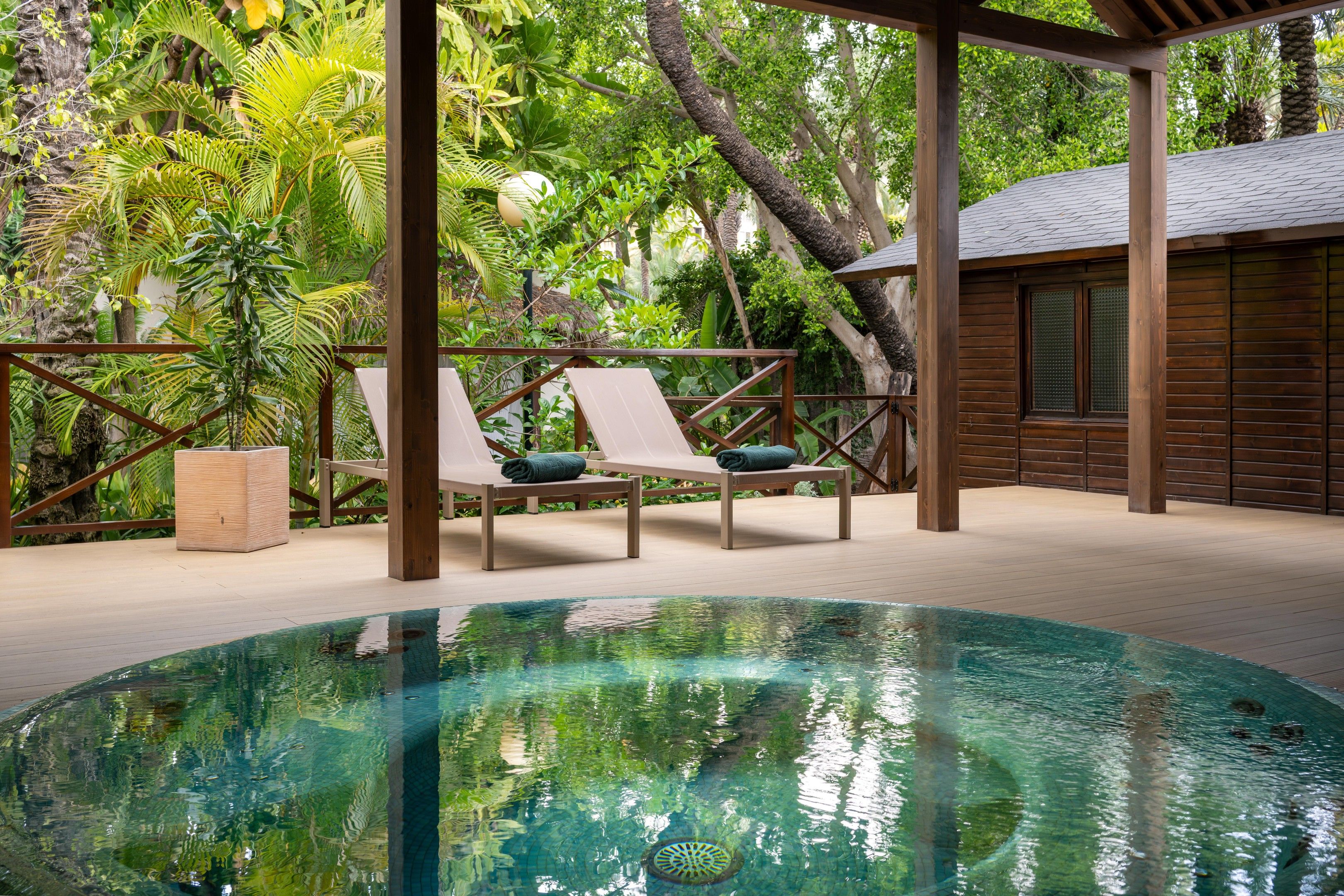 Un área de piscina con dos sillones de descanso, una terraza de madera y exuberante vegetación tropical.