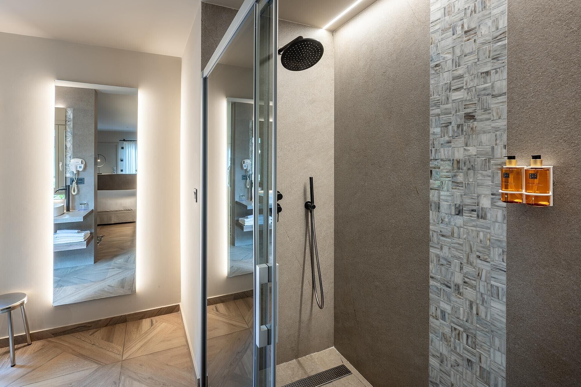 A modern bathroom with a glass shower enclosure, a marble-patterned wall, and a small seating area with a chair.