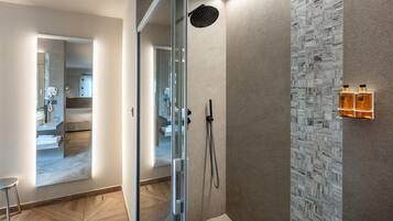 A modern bathroom with a glass shower enclosure, a marble-patterned wall, and a small seating area with a chair.