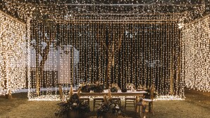 Un área de comedor al aire libre con una mesa dispuesta para una comida, rodeada de luces colgantes y centros de mesa florales.