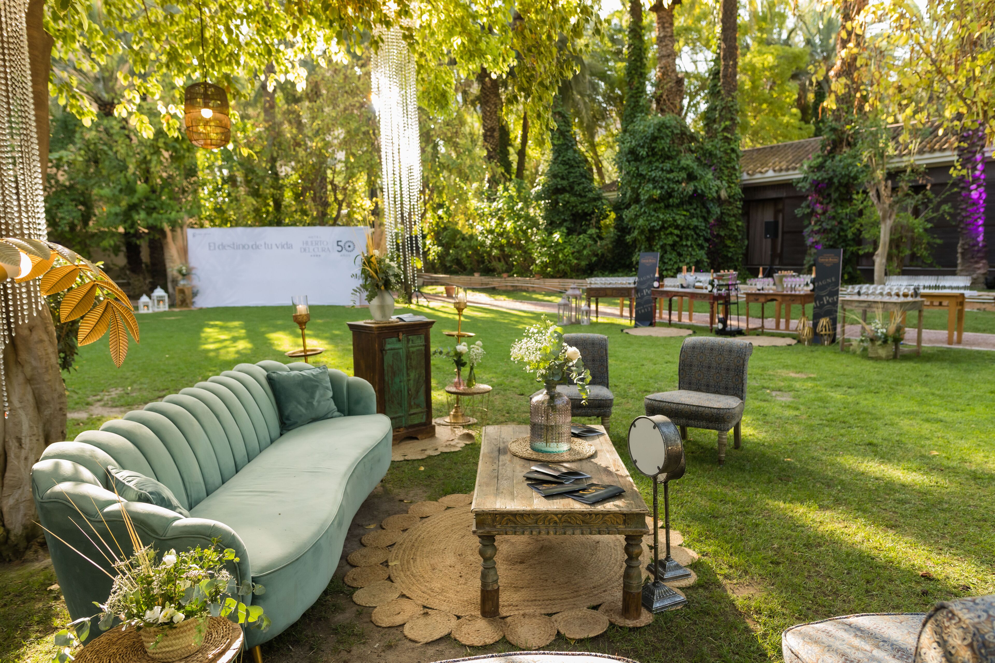 An outdoor seating area with a green sofa, a wooden table, and chairs, surrounded by greenery and a sign in the background.
