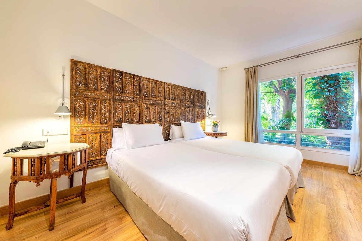Une chambre d’hôtel avec un grand lit, une tête de lit en bois, une table de chevet avec une lampe et une fenêtre donnant sur un paysage verdoyant.