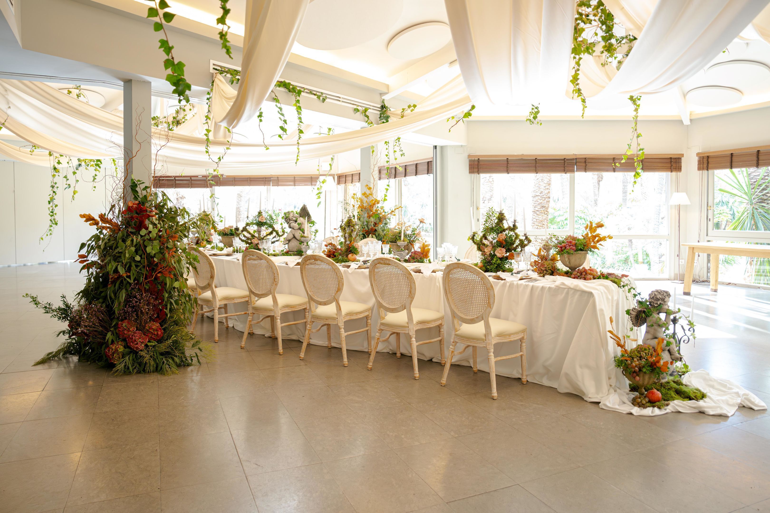 A banquet hall with a long table set for a formal event, surrounded by chairs and decorated with floral arrangements.