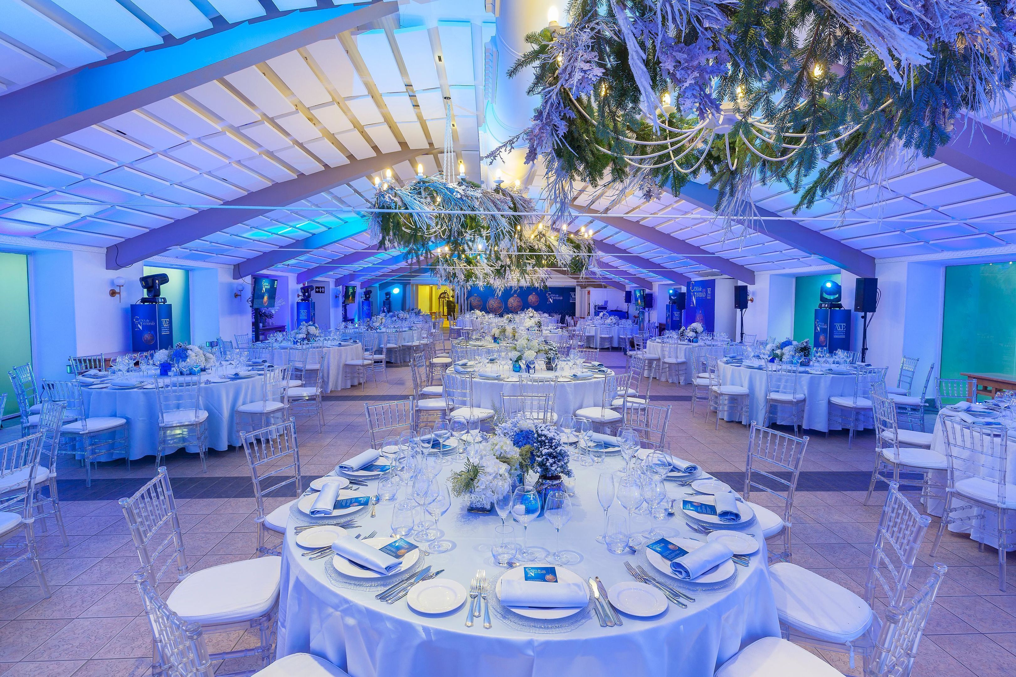 A large indoor venue with multiple round tables set for events, featuring clear chairs and white tablecloths.