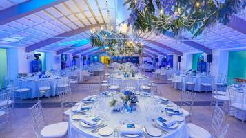 A large indoor venue with multiple round tables set for events, featuring clear chairs and white tablecloths.