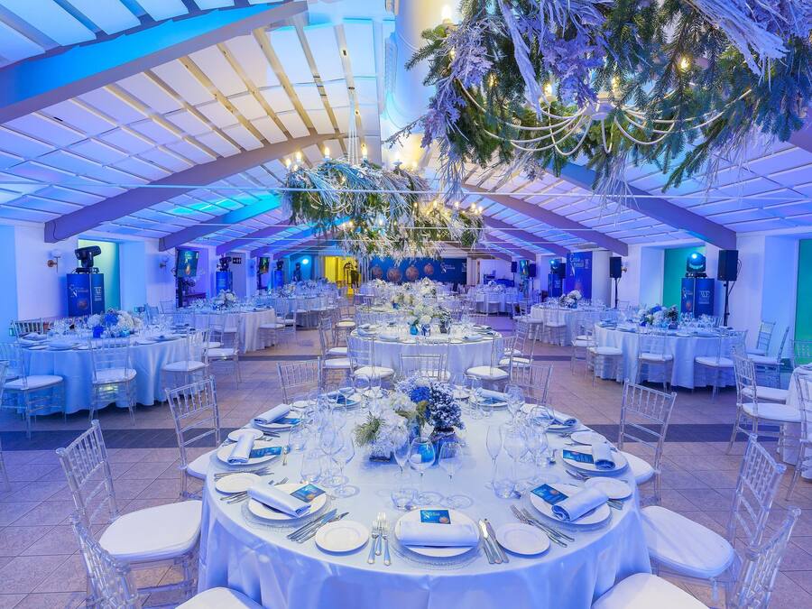 A large indoor venue with multiple round tables set for events, featuring clear chairs and white tablecloths.