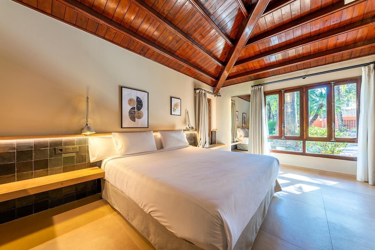 Une chambre spacieuse avec un grand lit, des poutres apparentes en bois au plafond, un mur recouvert de carreaux et une fenêtre donnant sur un paysage verdoyant.
