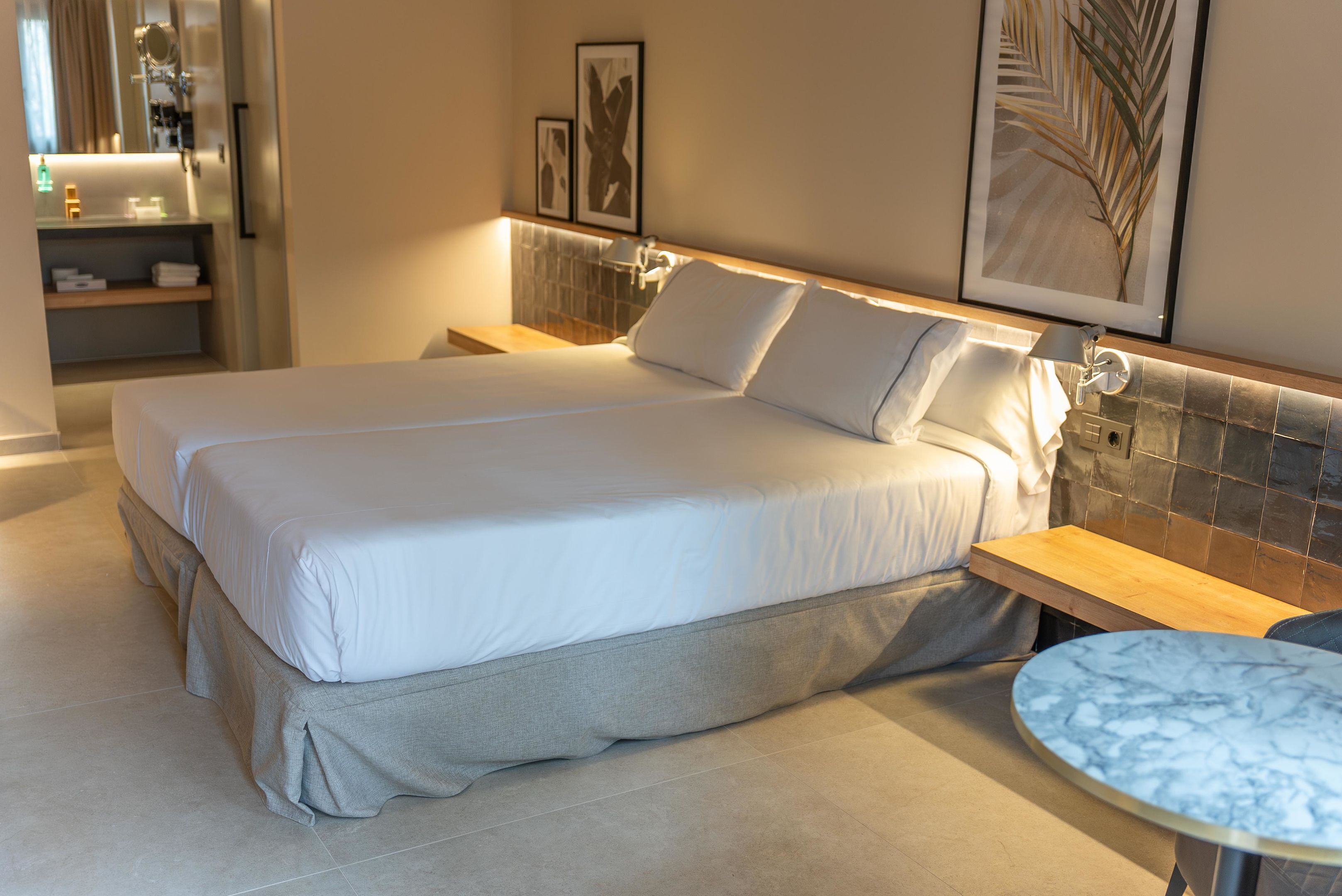 Una cama doble con falda gris, una mesita de noche, una mesita redonda de mármol y obras de arte en las paredes.