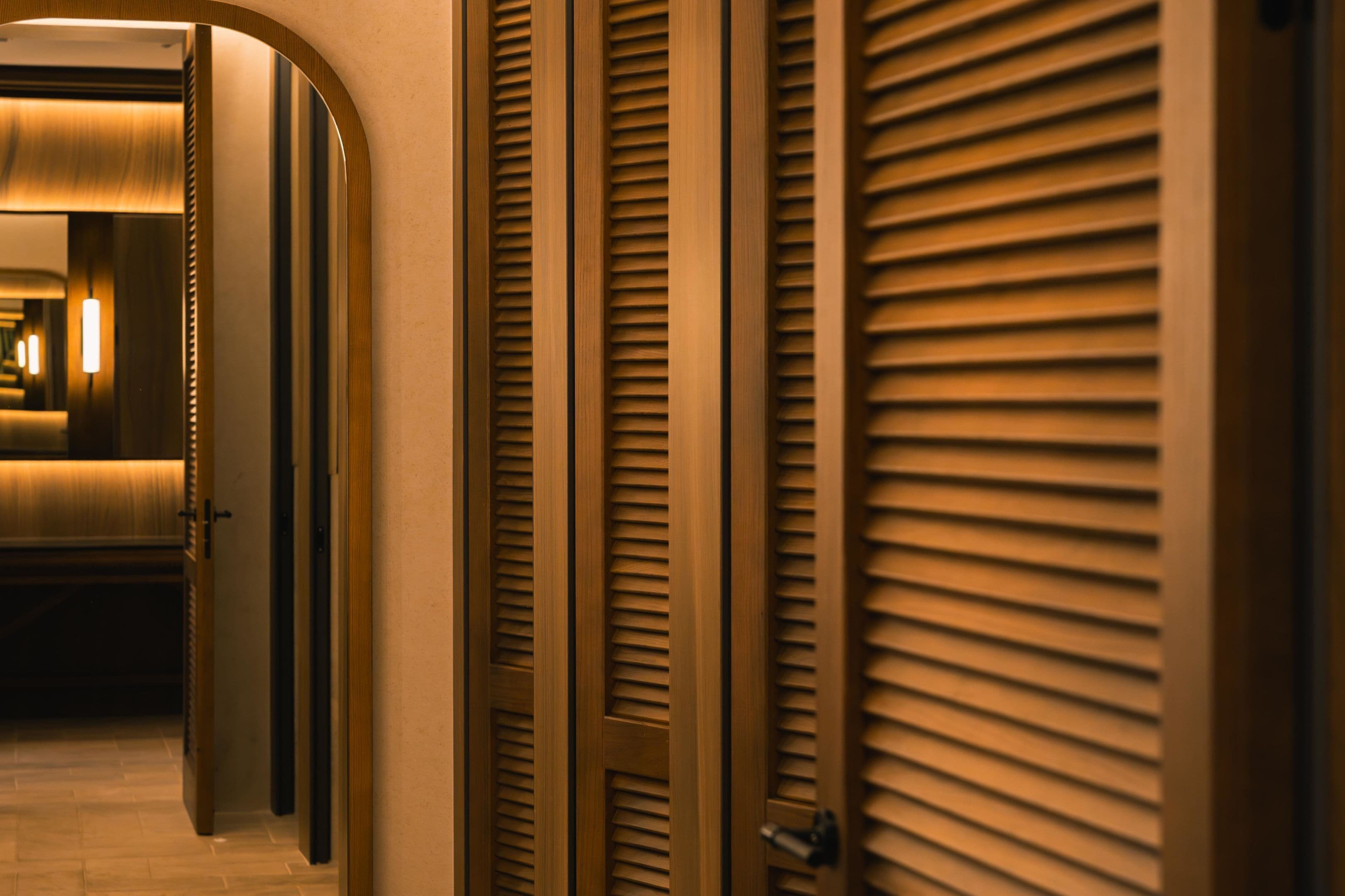 A corridor with wooden doors and a mirrored wall reflecting a lit interior.
