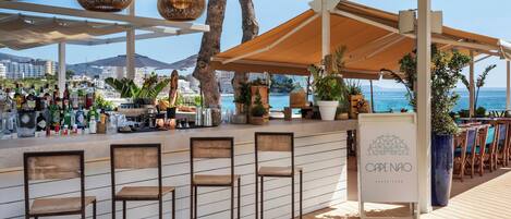 A beachside bar area with wooden chairs, a counter with bottles, and a sign reading "CAPE NAO BEACH CLUB".