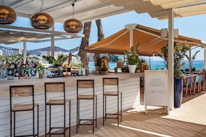 A beachside bar area with wooden chairs, a counter with bottles, and a sign reading "CAPE NAO BEACH CLUB". - Melia South Beach (Calvia)