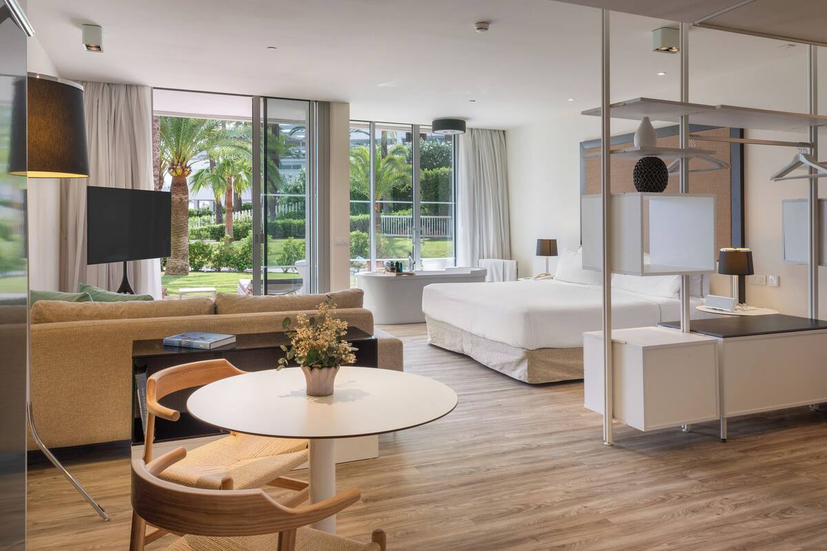Une chambre d’hôtel moderne dotée d’un grand lit, d’un canapé, d’une table à manger avec des chaises, d’une télévision et d’un balcon offrant une vue sur les palmiers.
