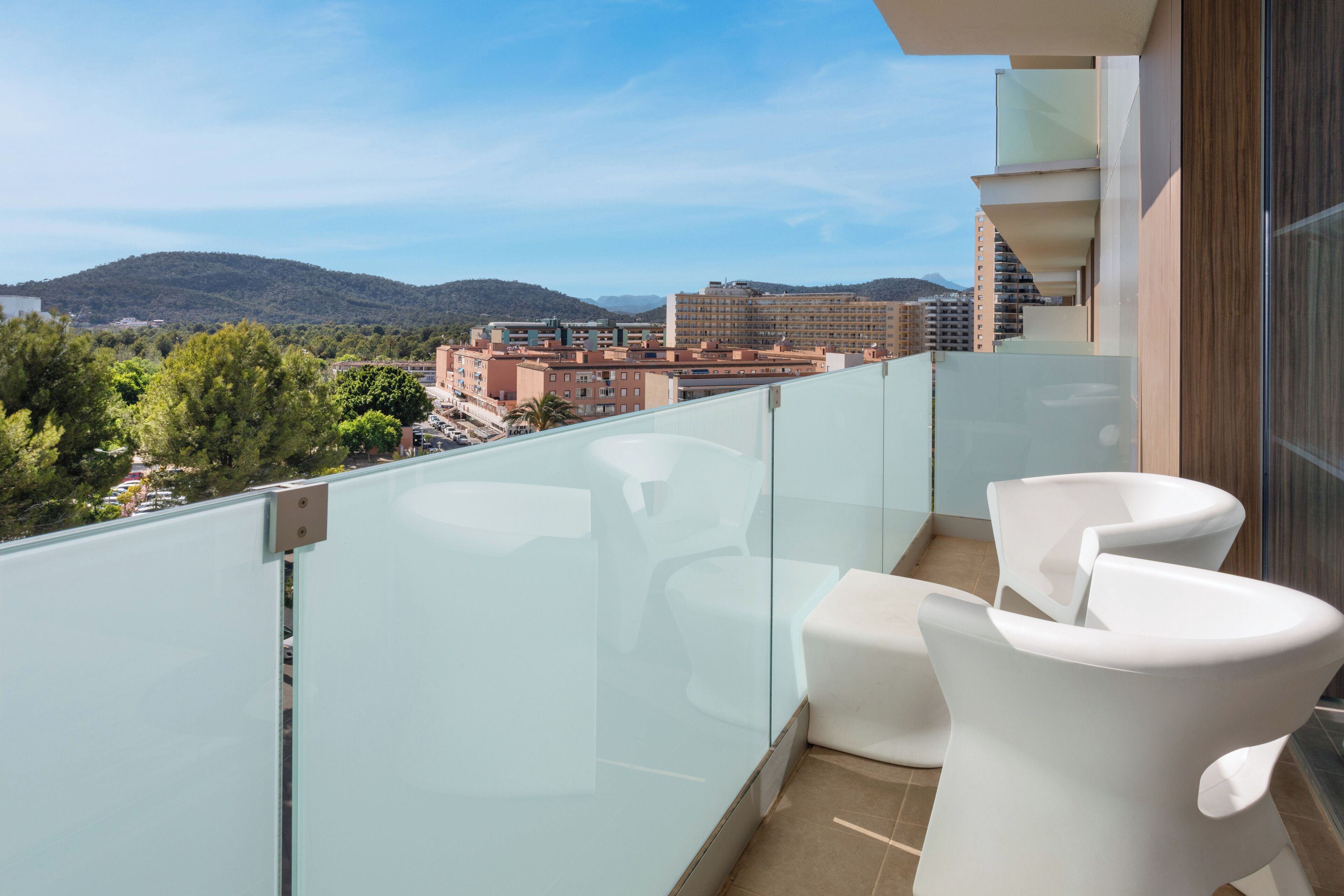 A balcony with a glass railing, white chairs, and a view of buildings and hills in the background.