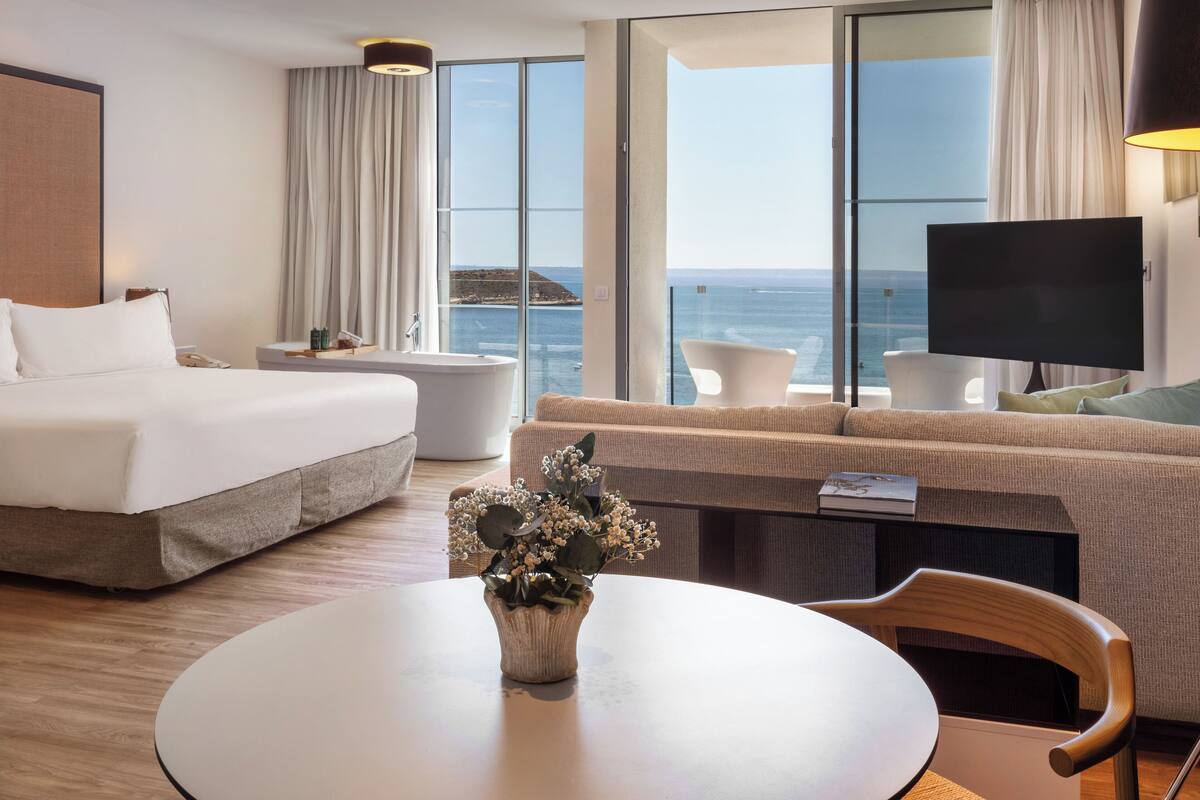 Une chambre d’hôtel avec un lit, une table ronde ornée d’un vase de fleurs, un canapé, une télévision et une vue sur l’océan.