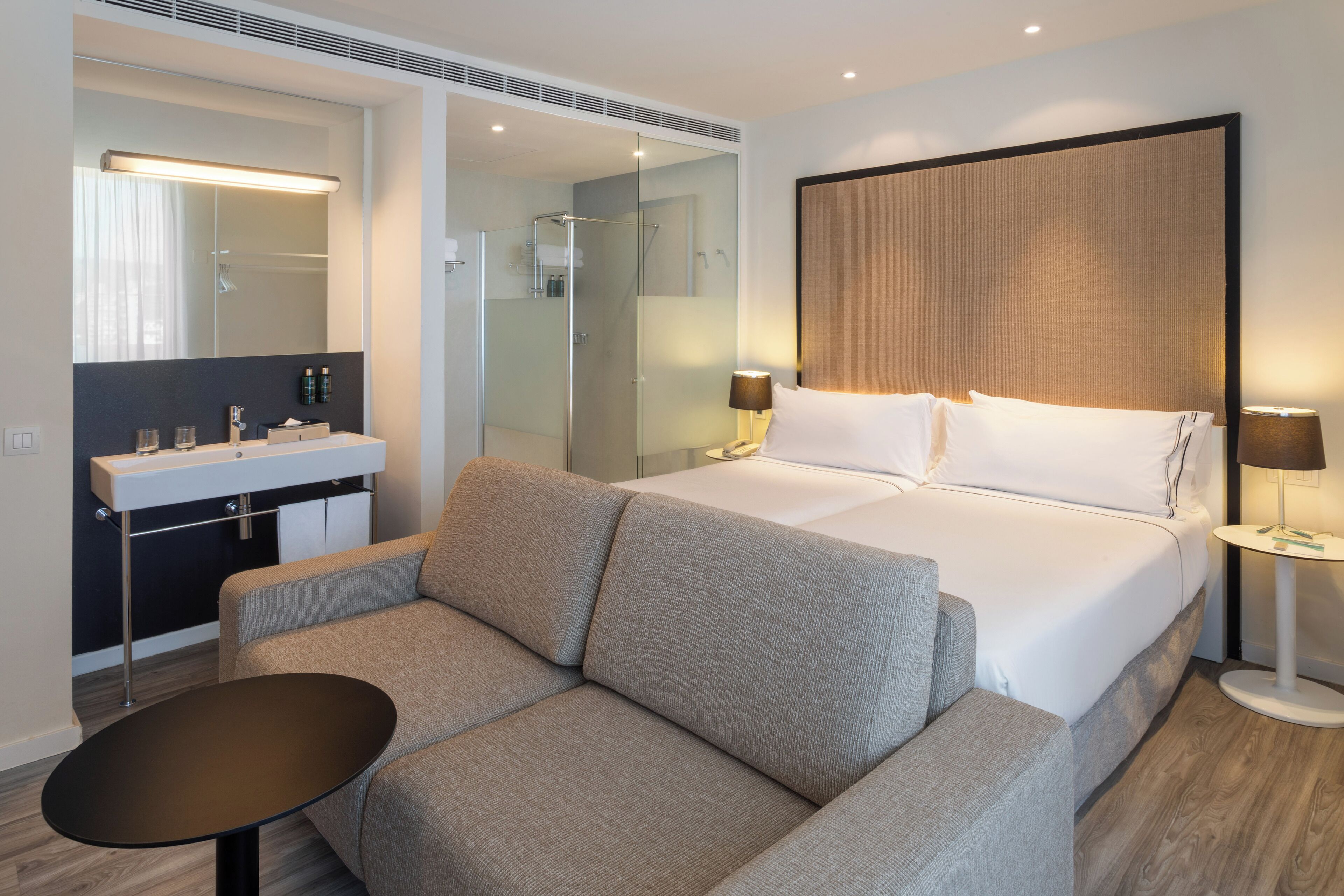 A modern hotel room with a bed, a sofa, a small table, a bathroom with a mirror, and a bedside lamp.