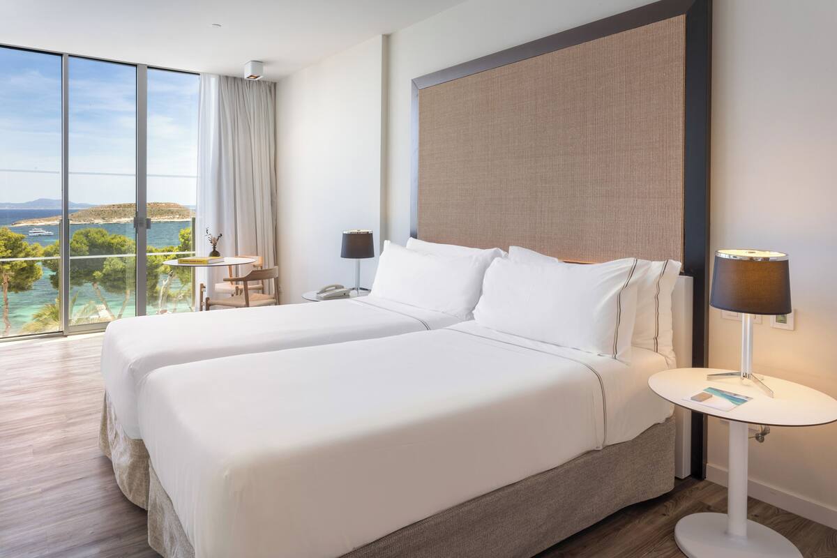 Une chambre d’hôtel avec deux lits, une vue sur la mer, une petite table avec une lampe et une grande fenêtre.