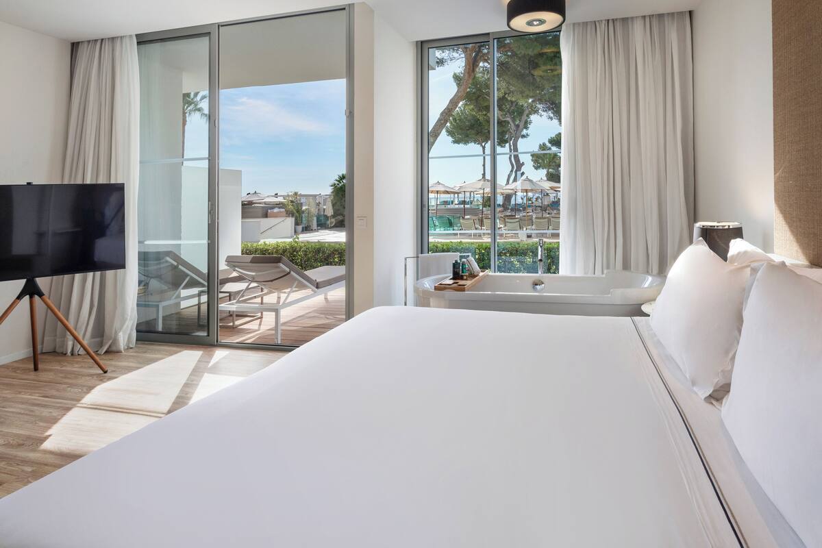 Une chambre d’hôtel moderne dotée d’un grand lit, d’un téléviseur sur un trépied, d’une baignoire et d’un balcon avec vue sur la piscine.