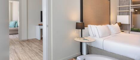 A hotel room with a large bed, a bedside table with a lamp, a wooden tray with toiletries, and a view of another room with a sofa and a window.