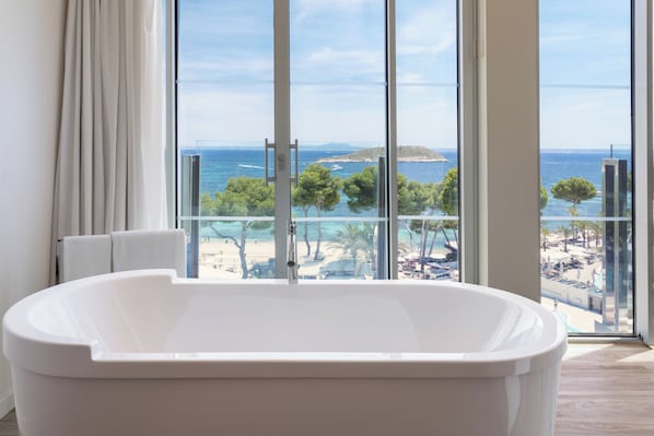 A modern bathroom with a large bathtub, large windows, and a view of a beach and sea. - Melia South Beach (Calvia)