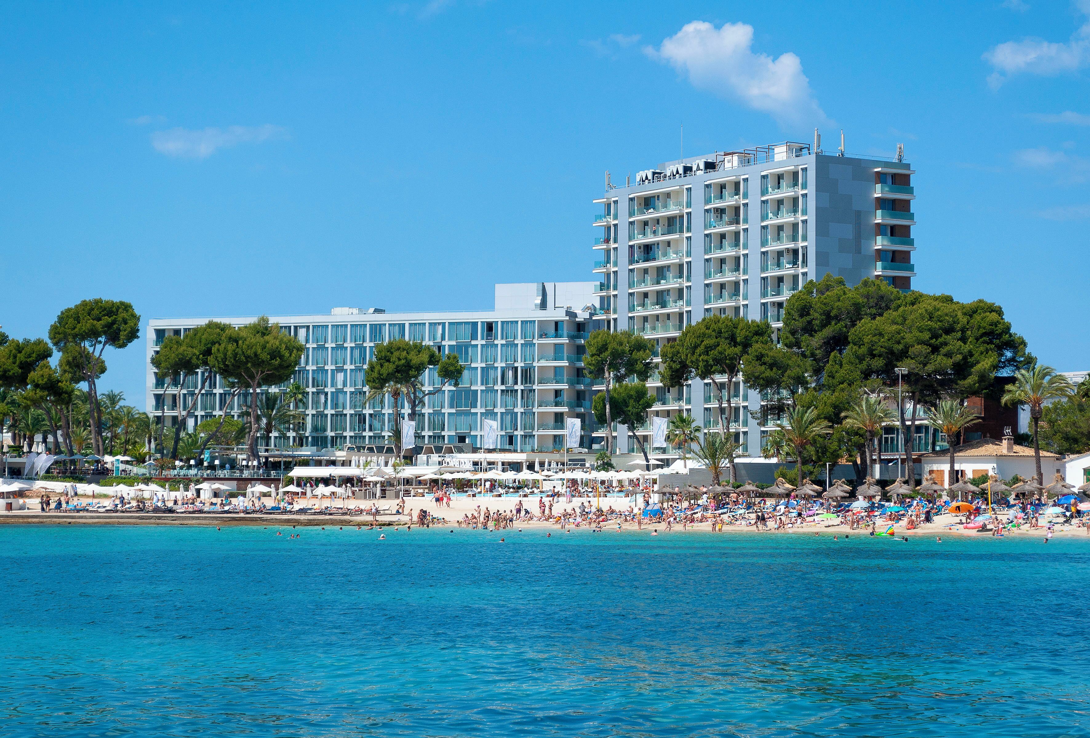 A hotel with multiple floors, a beach in the foreground, and palm trees.