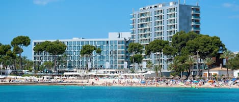 A hotel with multiple floors, a beach in the foreground, and palm trees.