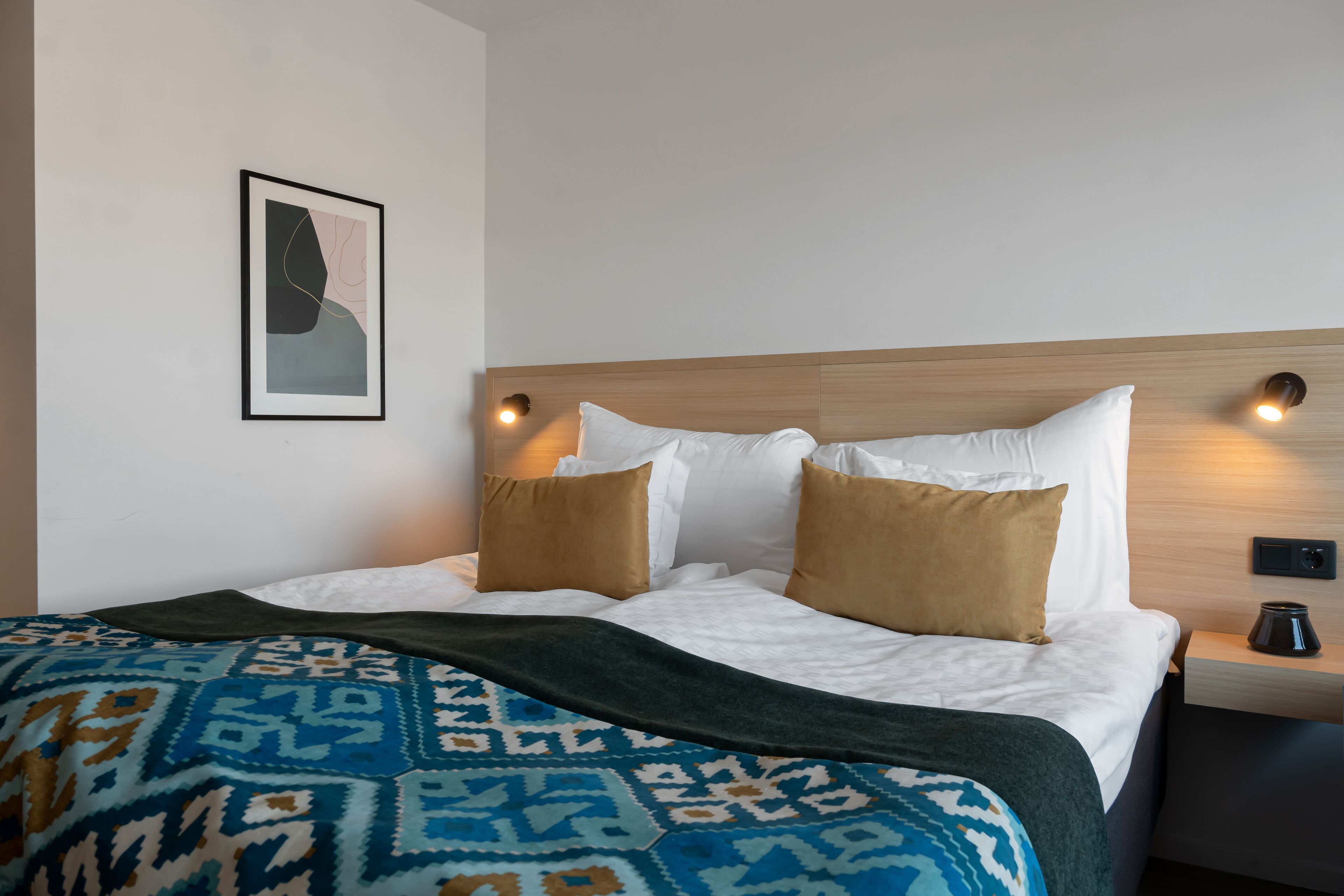 A hotel room with a bed, a nightstand, a wall-mounted lamp, and a framed picture.