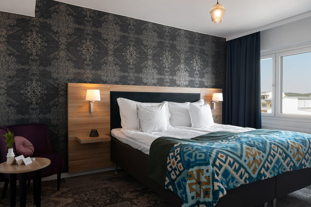 Hotellihuone, jossa on kuviollinen tapetti, puinen pääty, valkoisilla lakanoilla pedattu sänky, yöpöytä lampulla, pieni pöytä, jossa on kasvi, ja ikkunasta avautuva näkymä ulos.