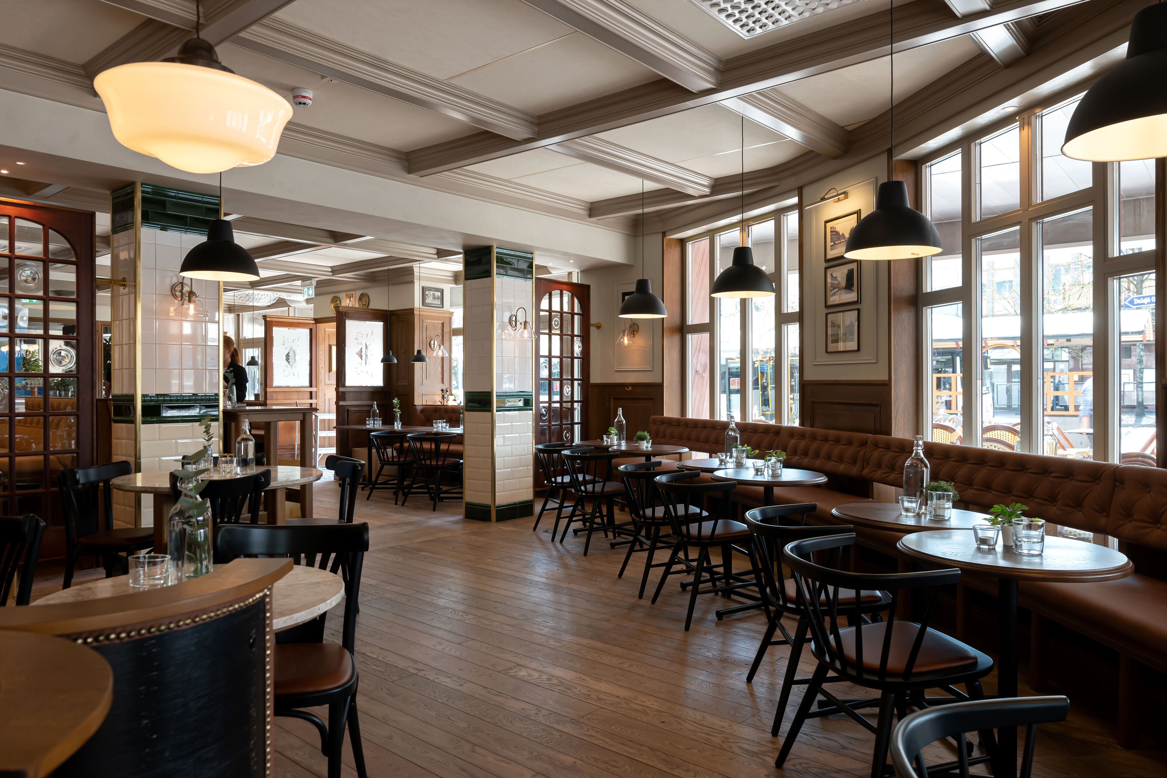 A restaurant interior with wooden floors, round tables, and black chairs. There are large windows allowing natural light and views of the street.
