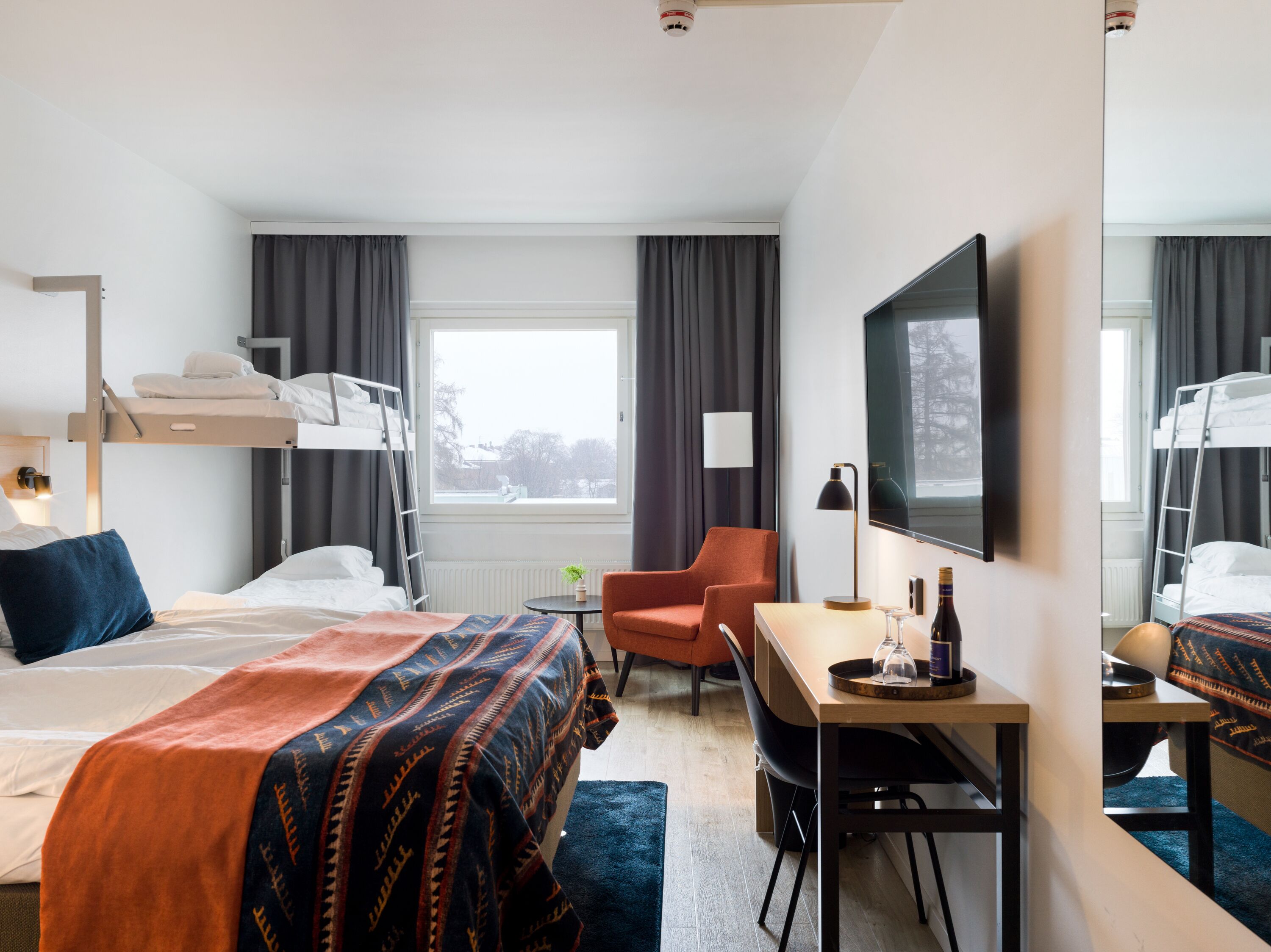 A hotel room with a bunk bed, a desk with a chair, a TV, and a window with curtains.