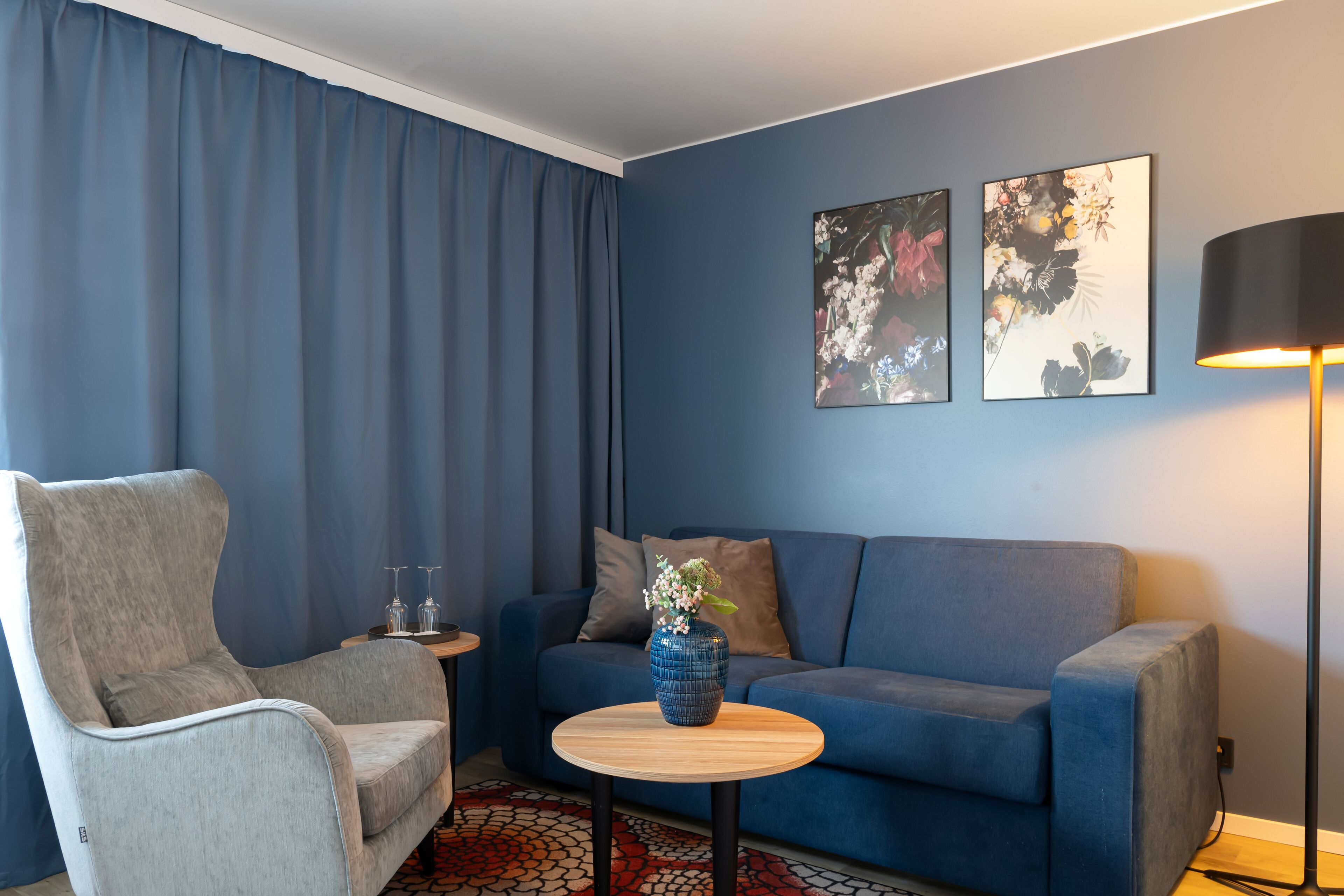 Una sala de estar moderna con un sillón gris, un sofá azul, una pequeña mesa redonda de madera con un jarrón, y dos cuadros florales colgados en la pared.