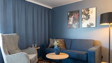 Una sala de estar moderna con un sillón gris, un sofá azul, una pequeña mesa redonda de madera con un jarrón, y dos cuadros florales colgados en la pared.