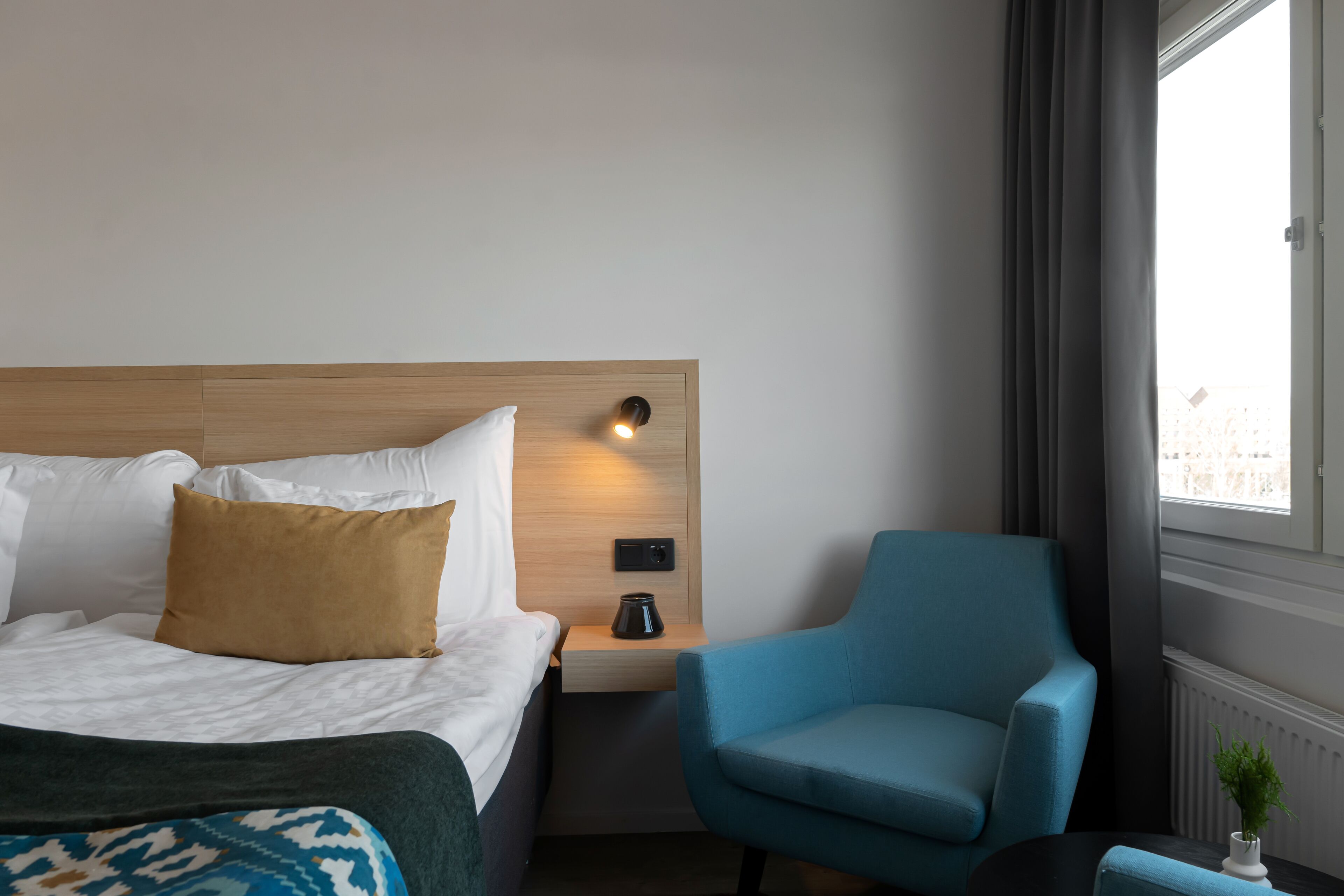 Habitación de hotel con cama, mesita de noche, lámpara, silla y ventana con cortinas.