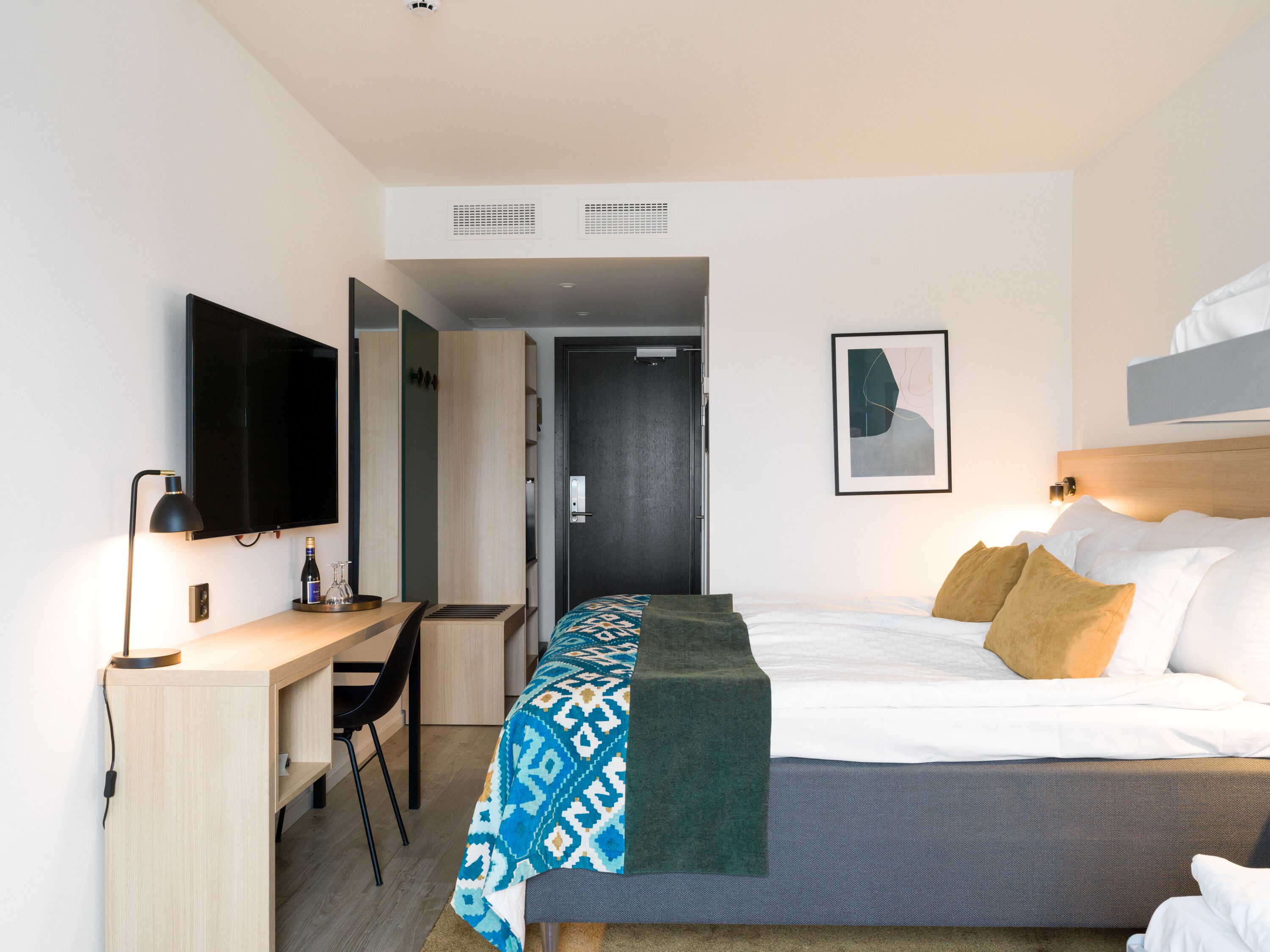 A modern hotel room with a bed, a desk, a chair, a TV, and a door.