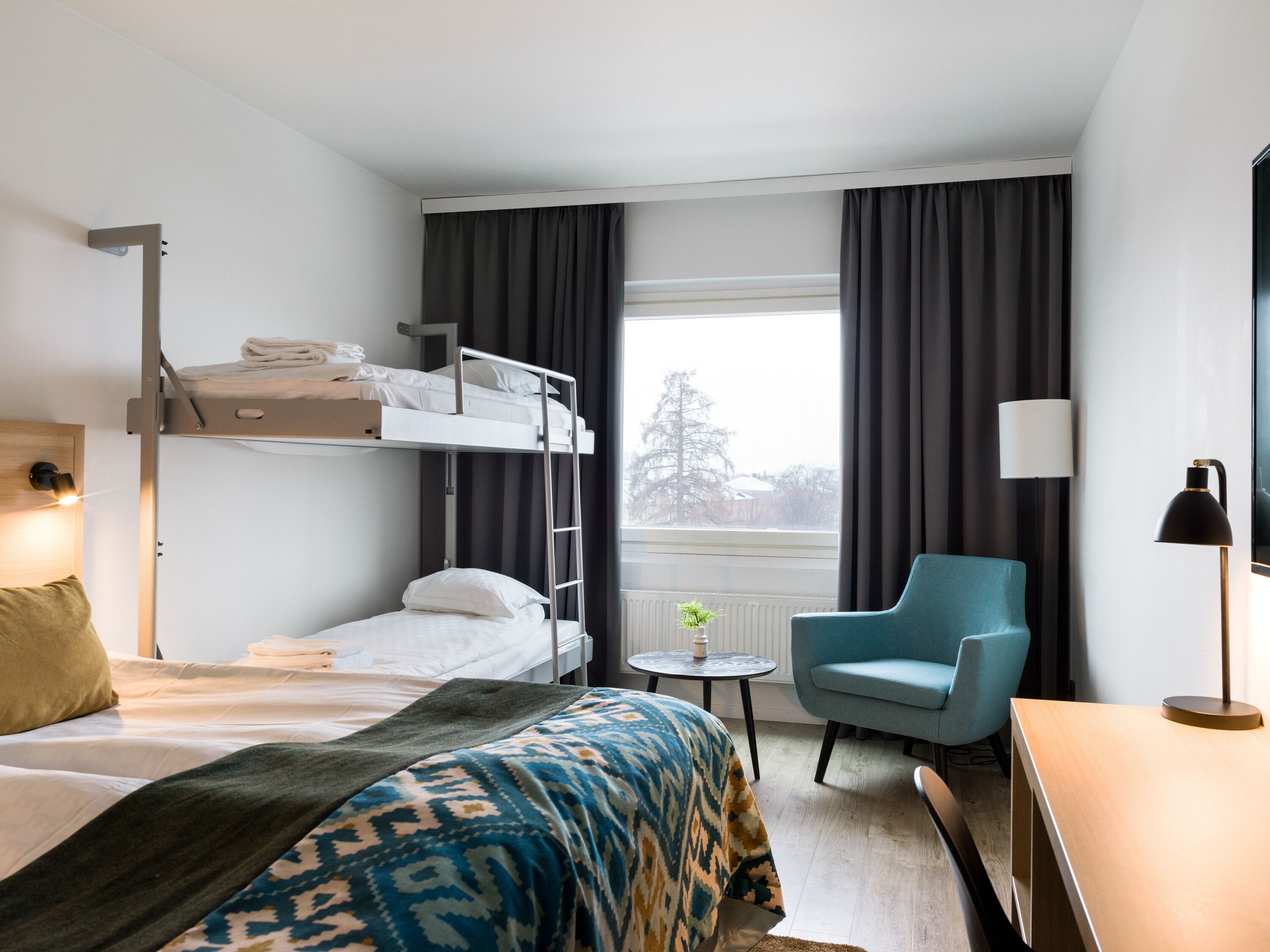 A hotel room with bunk beds, a single bed, a small round table, a blue armchair, a desk, and a lamp.