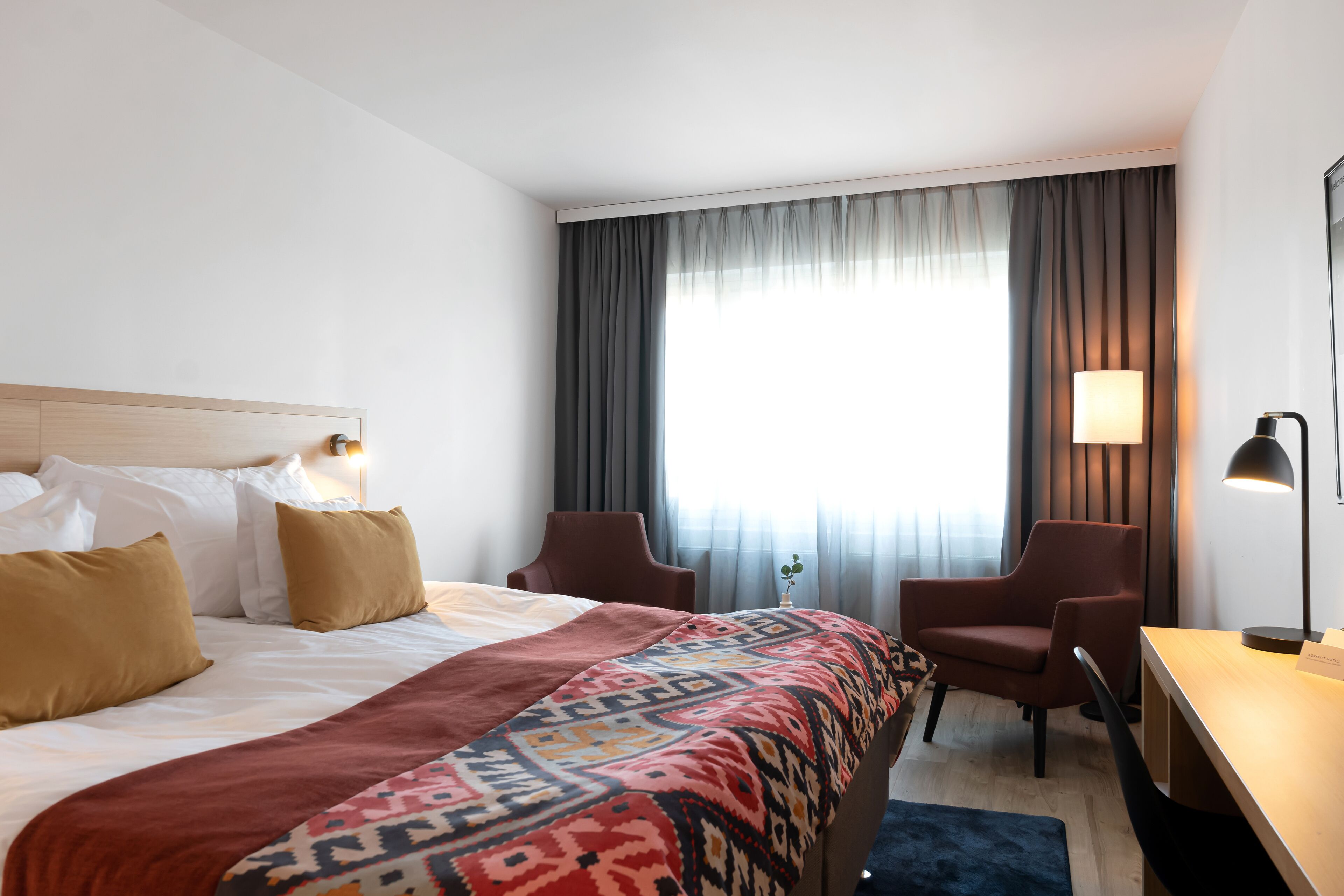 A hotel room with a bed, two armchairs, a desk, and a lamp.
