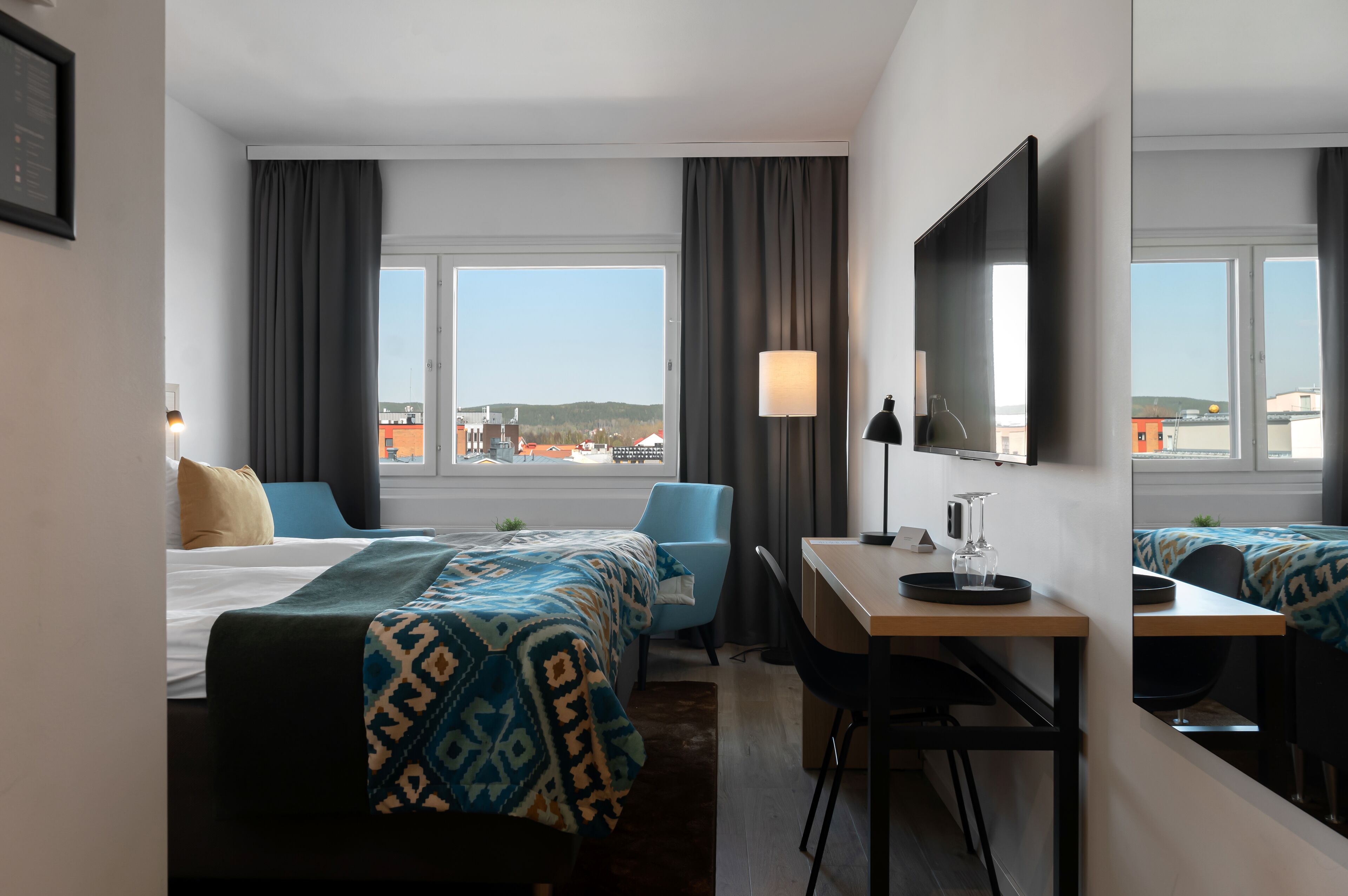 Habitación de hotel con cama, escritorio, silla, lámpara y un ventanal con vistas a edificios y colinas.