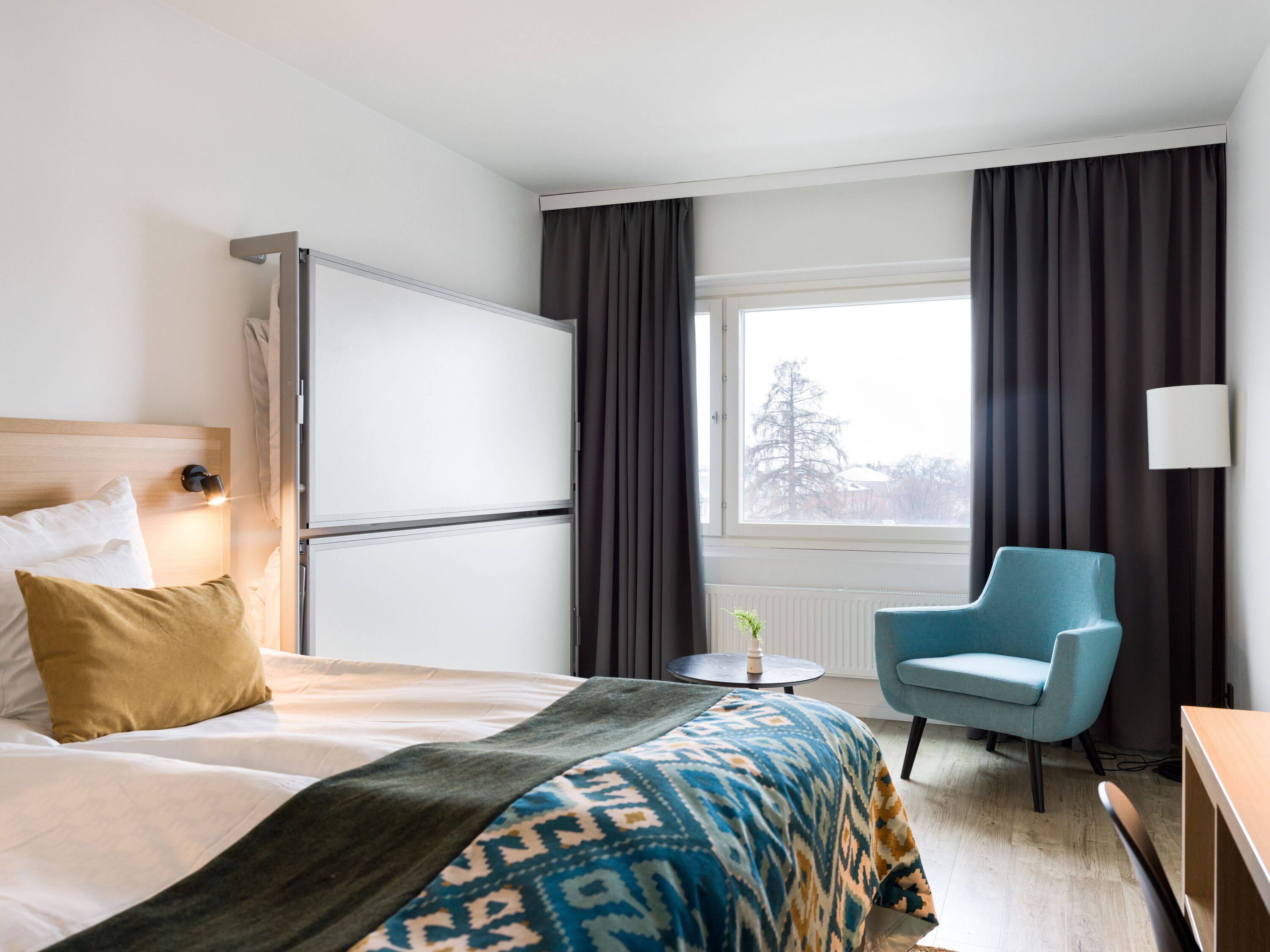 Habitación de hotel con cama, escritorio, silla, ventana con cortinas y vista a árboles.